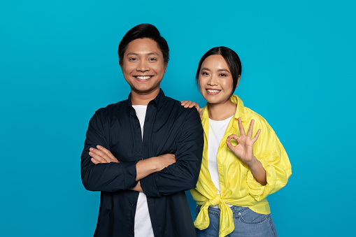 Confident millennial Asian millennial man with crossed arms and smiling woman showing an 'okay' sign, both looking pleased and content, set against a vibrant turquoise background,studio