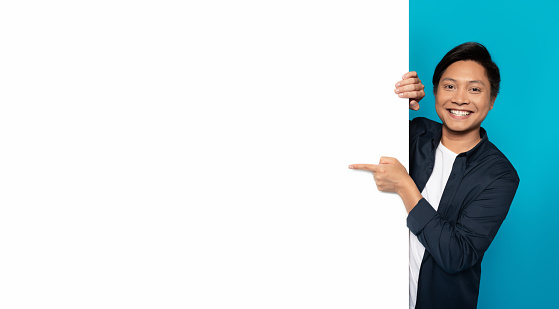 An Asian man with a cheerful smile is playfully peeking from behind a blank white vertical banner, pointing towards it, perfect for customizable content on a turquoise background