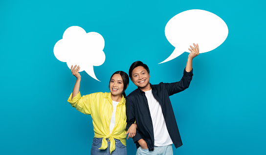 An exuberant Asian couple joyfully holds up speech bubbles, symbolizing lively conversation or the sharing of ideas, against a vivid turquoise backdrop with plenty of space for text