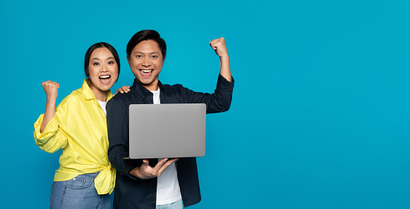 Overjoyed millennial Asian couple triumphantly raising fists while holding a laptop, signifying success or victory, against a vibrant turquoise blue background, studio