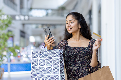 Smiling young Indian girl shopping in shopping center, standing inside with bags in hand, holding credit card and looking at mobile phone screen.