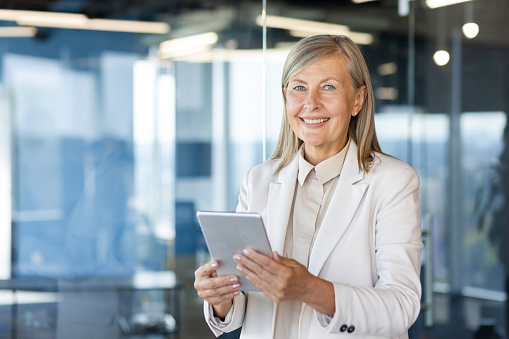 Close-up portrait of a senior smiling business woman standing in the office, holding a tablet in her hands and looking confidently into the camera.