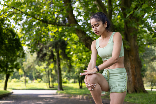 Indian young girl standing in a park in a tracksuit and holding her hands on her knee, feeling a lot of pain, sprained her muscles after running and doing sports.