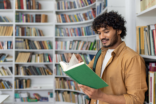 An adult male stands in a well-lit library, immersed in reading a green book. Bookshelves filled with various books surround him, suggesting a quiet atmosphere for study and literature exploration.