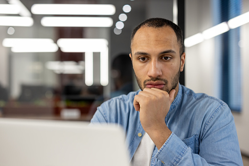 A close-up portrait of a young African American man sitting in the office in front of a laptop screen, intently and focused on work, a project, thinking about a solution to a problem.