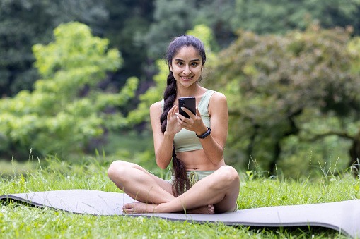 Young Indian woman in a tracksuit uses her phone while sitting on a yoga mat in a serene park, embodying fitness and relaxation.