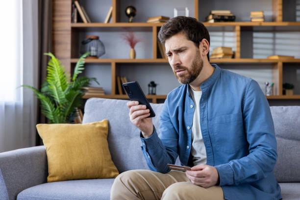 istock Concerned man sitting on couch looking at phone and card 2105330532