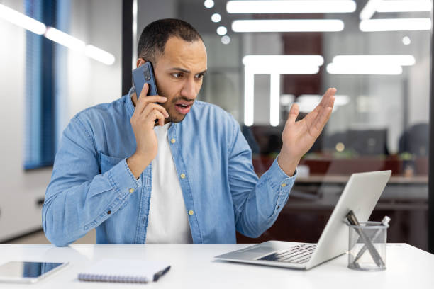 istock A man is talking on his cell phone while sitting at a desk with a laptop 2105358312