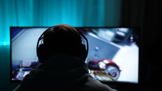 Back view of unrecognizable gamer male in headphones playing first-person shooter online video game on powerful personal computer sitting at desk in dark room