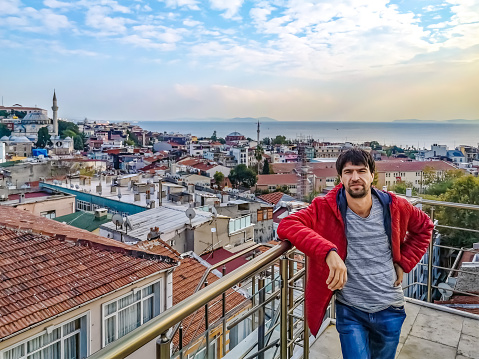 Young man poses against the backdrop of the red tiled roofs of Istanbul and the Marmara Sea skyline, Turkey. Adult guy stands on the roof of a residential building in a Turkish city