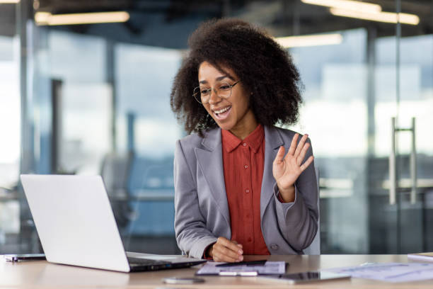 istock Friendly female making hi gesture while sitting by desktop with modern laptop at personal workspace. Hr manager starting interview with candidate for employment and creating positive atmosphere 2105474225