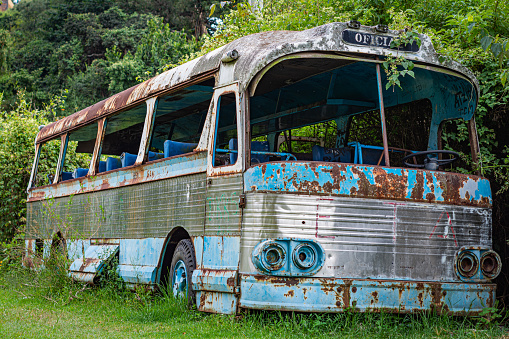 A rusted old bus is sitting in a grassy field. The bus is blue and white and has a faded orange lettering on the side. The bus appears to be abandoned and has been left to rust in the grass
