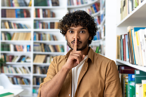 A young adult male with curly hair placing a finger on his lips, signaling quietness amidst a backdrop of bookshelves filled with various books. He portrays a sense of silence and focus, typical of a library or study environment.