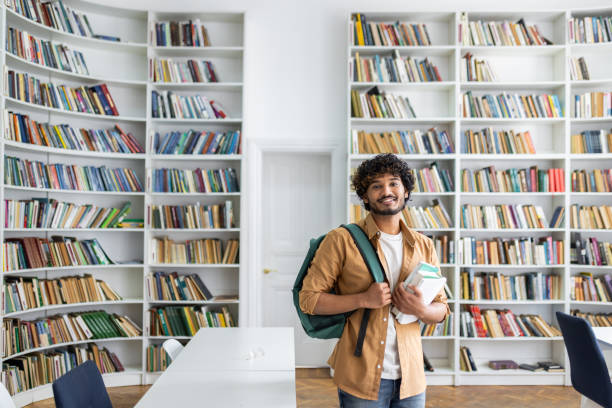 istock Smiling young man with backpack holding books in a library 2105531102