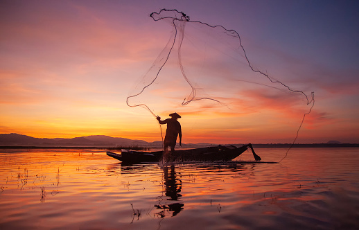 Fisherman casting his net on during sunset. Silhouette Asian fisherman on wooden boat casting a net for freshwater fish. Thailand culture.