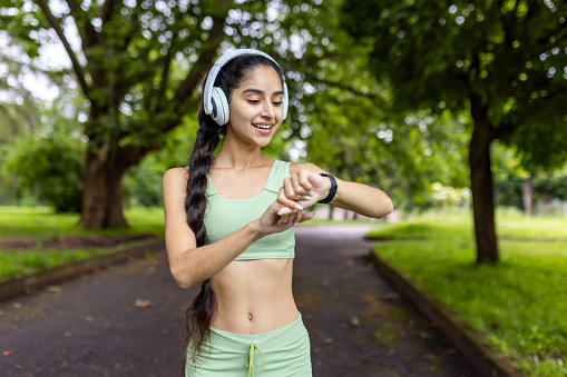 A young Indian woman in a green tracksuit is checking her fitness tracker while exercising in a lush park with headphones.