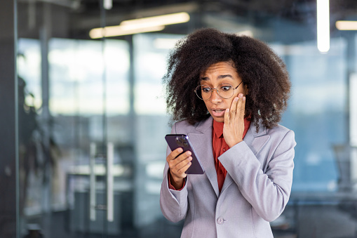 Shocked young African American woman standing in office space and looking at phone screen in surprise, holding hand to face.