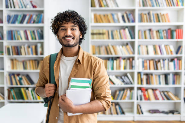 istock Happy unshaved man wearing backpack and holding stack of books while smiling at camera at bookstore. Optimistic classman making purchases for beginning of new academic semester in college 2105591779
