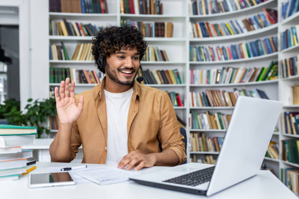 istock Friendly man waving in virtual meeting with laptop and headset at library 2105593821