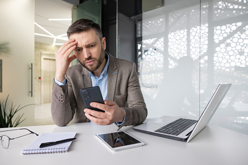 Upset frustrated man holding phone inside office, sad businessman reading bad news online, boss experienced financier accountant unhappy with workplace achievement results.