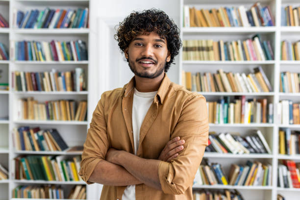 istock Confident diverse man in casual outfit looking at camera with arms folded on chest on background of closet with books. Intelligent male posing in library of university campus with happy expression 2105608420