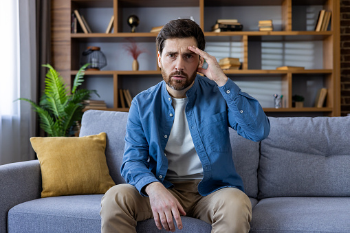 A pensive adult male with a beard sits on a sofa, appearing stressed or troubled, surrounded by a cozy home environment.
