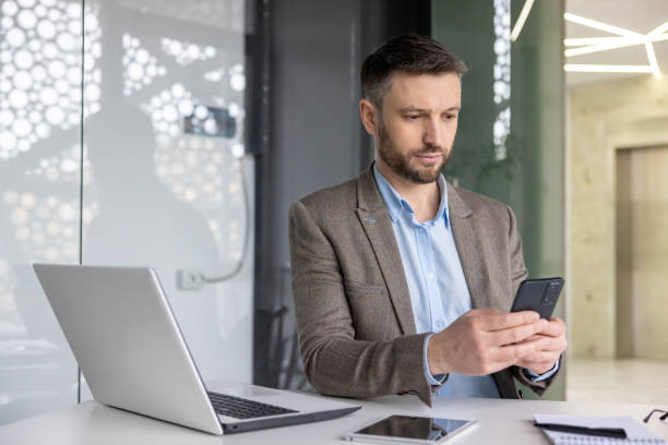 istock Serious concentrated mature businessman thinking reading online social networks, experienced mature man holding phone using smartphone at workplace inside office with laptop 2105692969