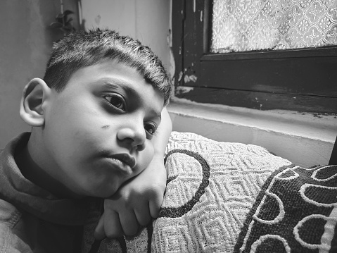 Black and white close-up portrait of sad, serene indian boy looking out through the window and contemplating with a blank expression at home