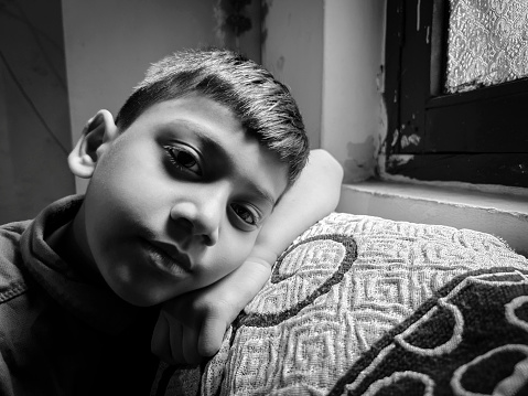 Black and white close-up portrait of sad, stress indian boy sitting alone on the couch near the window and looking at the camera with a blank expression and contemplating  at home