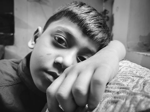 Black and white close-up portrait of sad, stress indian boy sitting alone on the couch near the window and looking at the camera with a blank expression and contemplating  at home