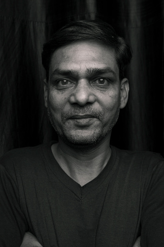 Black and white portrait of mature man standing against a black background and looking at the camera with smile.