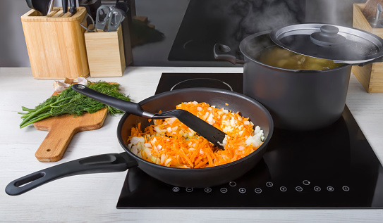 A frying pan with carrot and onion roasting for soup and a saucepan with boiling broth on an electric glass-ceramic hob in a modern kitchen