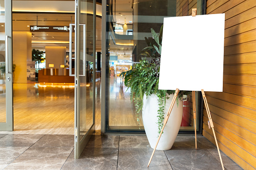 A photo of a blank white canvas on an artist's easel in a studio. The canvas is in a vertical orientation and is slightly off-center in the frame. The easel is made of wood and has three legs.