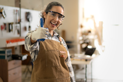 Portrait of Latin carpenter standing with thumb up in workshop smiling. Hispanic woodworker woman happy working at furniture carpentry woodshop. Beautiful small business owner in woodwork industry