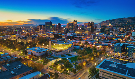 Downtown Salt Lake City skyline aerial at dusk with a park and illuminated streets and autumn colored trees in the foreground.
