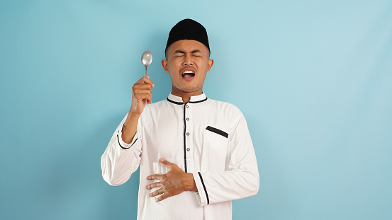 Asian muslim man hungry expression and holding spoon on blue light background