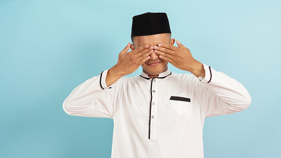 Muslim man covering eyes with hands, something that should not be seen during ramadan, isloated on blue light background