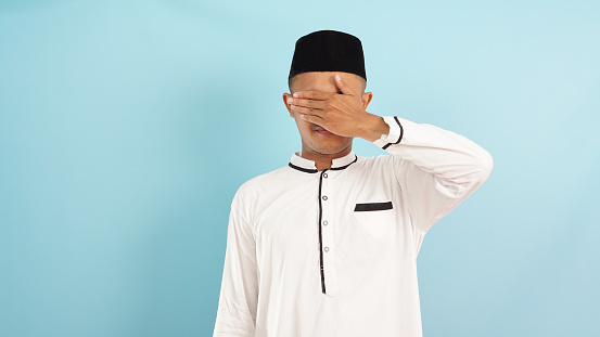Muslim man covering eyes with hands, something that should not be seen during ramadan, isloated on blue light background