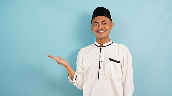 Asian  muslim man smiling showing something and presenting on his side, blue light background