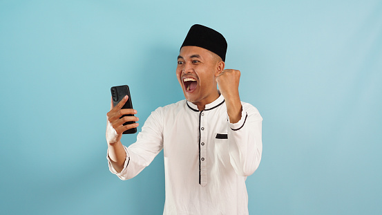 A muslim man excited get good news from phone, celebrating success on blue background