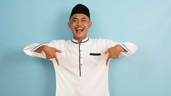 muslim man pointing down to free space with finger, happy expression for advertisement isolated on blue light background