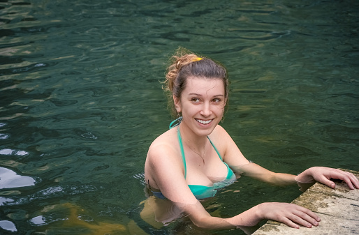 Portrait of young smiling attractive woman in bikini standing in water by wooden deck of lake