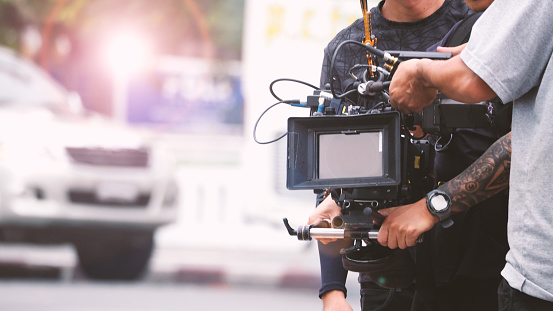 Blurred images of high definition video camera and lens on steady equipment support such as gimbal steady or stabilized shoulder rig and pan tilt shift head tripod for handheld filming a fast moving object in tv commercial production at outdoor location.