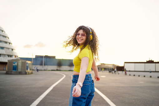 Young woman with curly hair turning to smile at camera while listening to music in the city