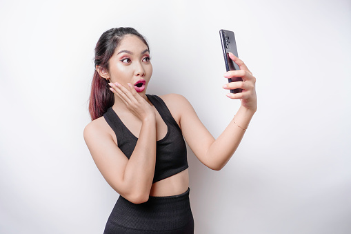Shocked sporty Asian woman wearing sportswear and holding her phone, isolated by white background