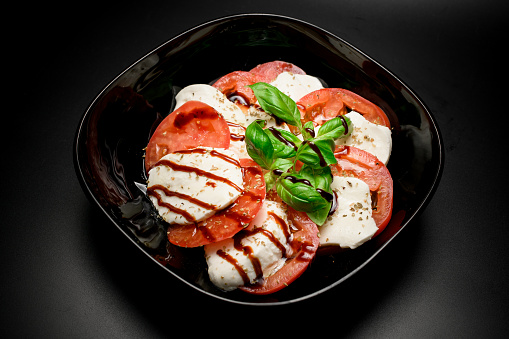 indulge in the timeless freshness of a caprese salad, elegantly presented in a black bowl, adorned with a flavorful finishing sauce.
