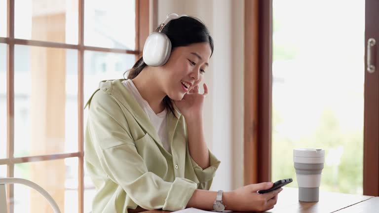 An Asian businesswoman in casual attire is working remotely at a cafe, selecting music on her smartphone, and using headphones to relax from work