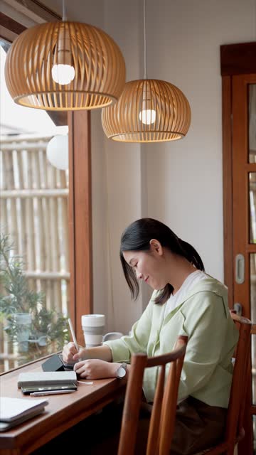 An Asian female entrepreneur working at a nearby cafÃ©, diligently taking notes on her tasks using a tablet, demonstrating focused remote work
