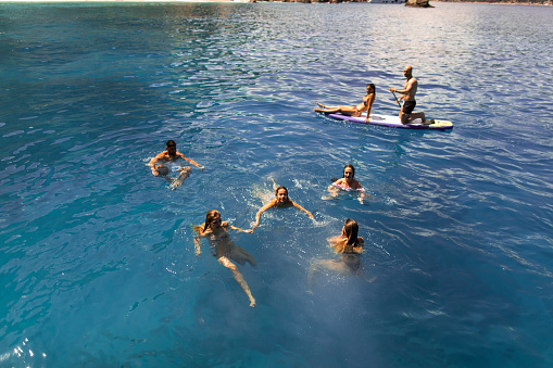 Group of young people swimming, paddleboarding and having fun next to the sailboat.  The sea is amazingly clear and blue.