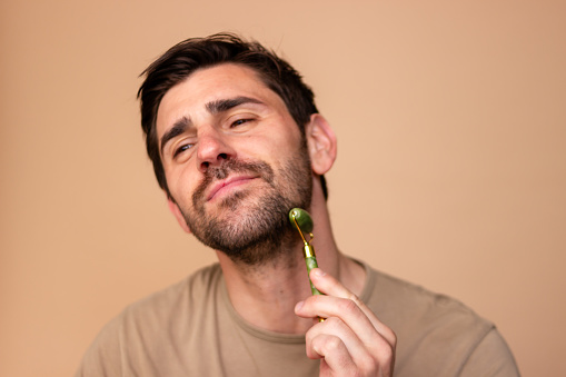 Content male uses a jade roller for a soothing skincare routine against a soft background